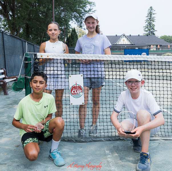 Four young tennis players pose beside a net on an outdoor court while holding rackets and a tournament cup after a match. The group portrait highlights teamwork and achievement often featured in sport photography for youth scholarship apps.