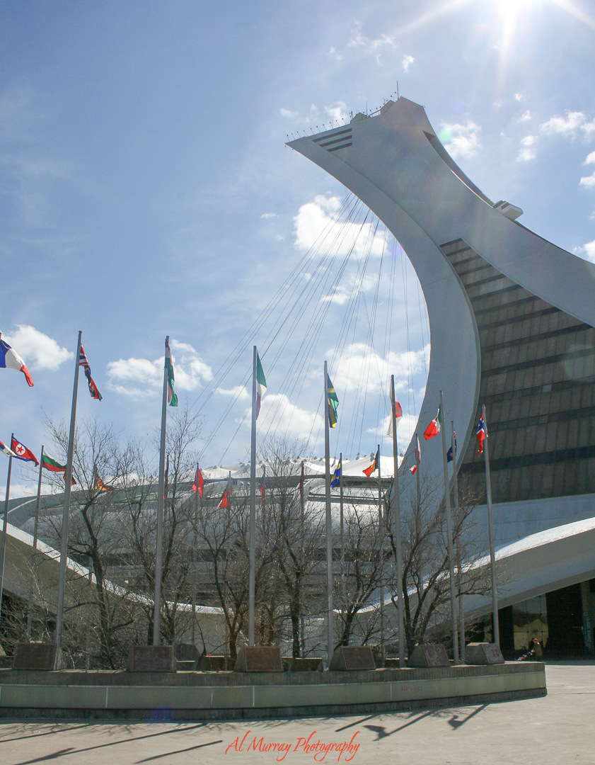 Montreal’s Olympic Stadium tower rises into a bright blue sky with sunlight flaring above and international flags lining the foreground plaza. The sweeping architecture and wide outdoor scene capture the perspective of a travel and landscape photographer exploring the city.