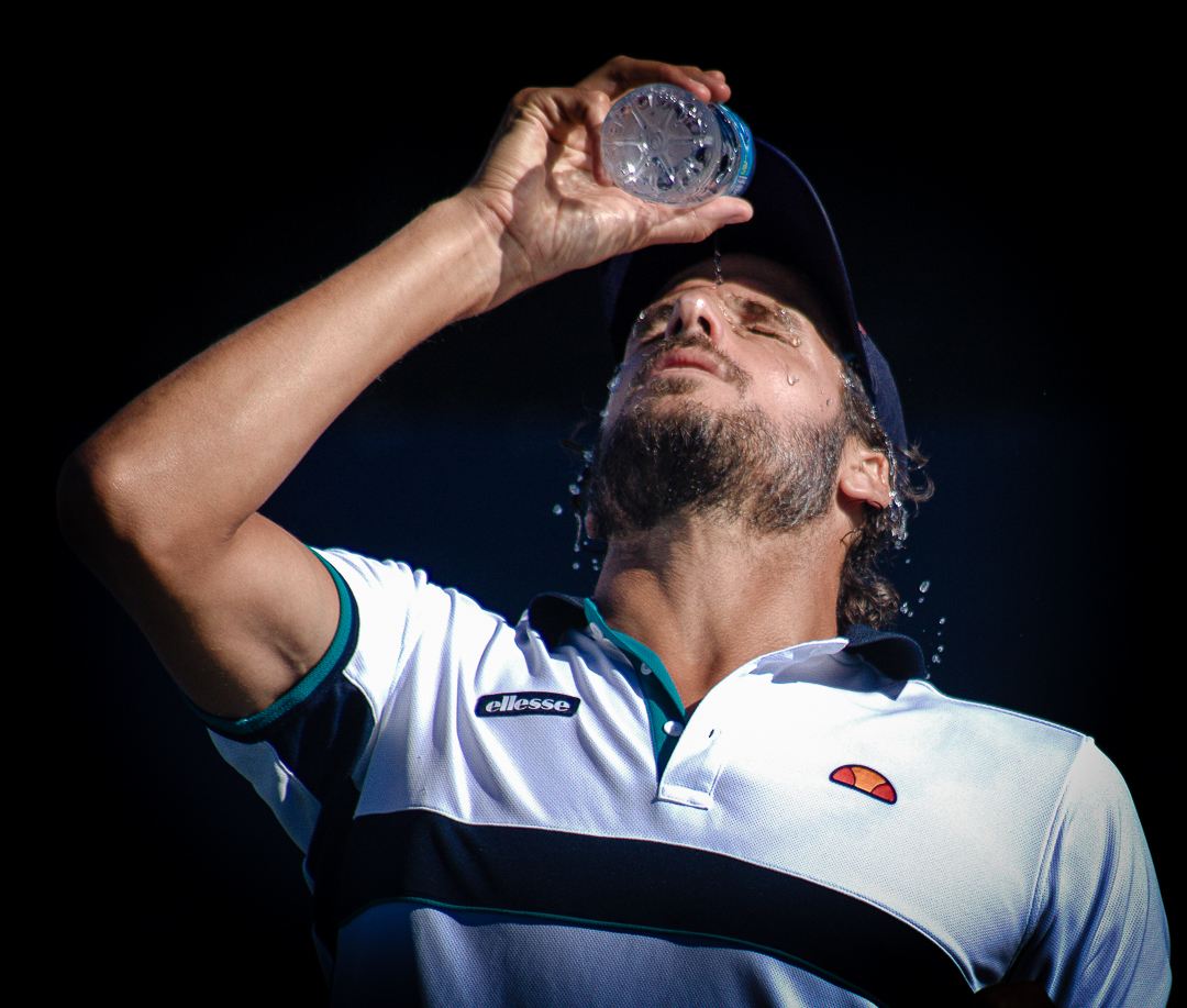Tennis player tilts his head back while pouring water over his face during a match break under bright stadium lighting. The close up composition captures the physical intensity and emotional moments often highlighted in international sport photography.