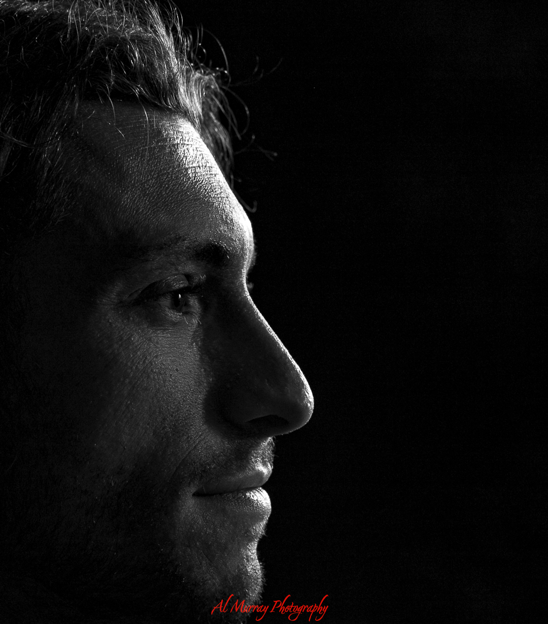 Black and white side profile portrait of a man softly lit against a dark background. Dramatic lighting highlights the texture of his skin and facial features, showcasing the style of a portrait photographer in Woodstock, Ontario.