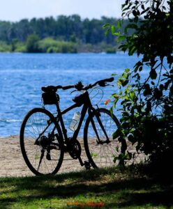 Black bicycle parked beside a calm blue lake with leafy branches casting shadows in the foreground and trees lining the distant shore. The peaceful outdoor setting captures the quiet pace and scenery often featured in travel photography.