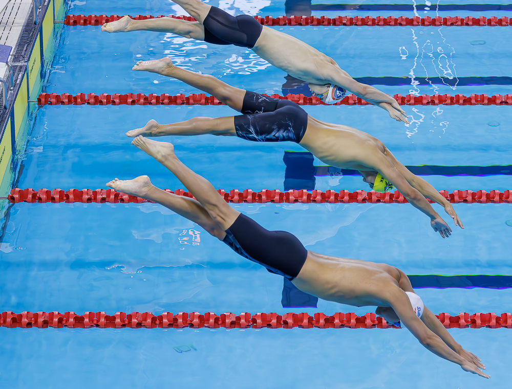 Competitive swimmers dive into a pool at the start of a race with their bodies stretched forward above bright blue water and red lane dividers. The synchronized motion and overhead perspective showcase the energy and timing often captured in amateur sport photography at local and national competitions.