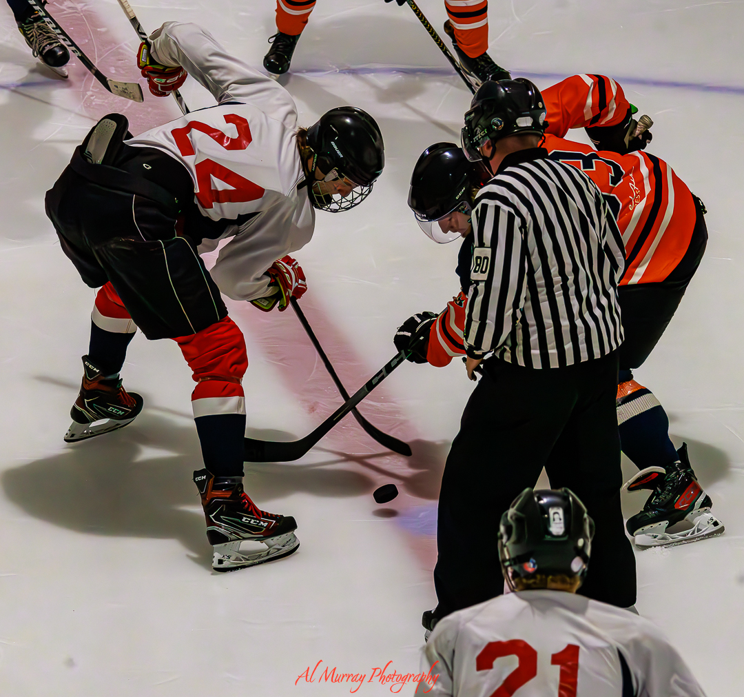 Hockey players in white and orange jerseys lean in for a faceoff while a referee drops the puck at center ice during a competitive game. The overhead angle and fast paced action capture the intensity and precision expected from a hockey photographer covering local and national events.