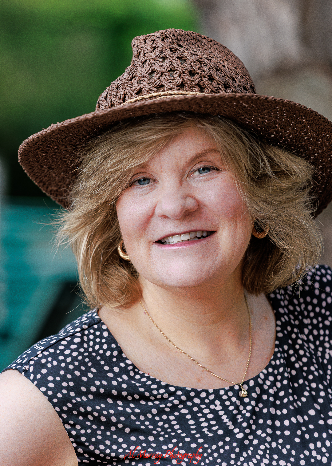 Smiling woman with short blonde hair wears a woven brown sunhat and patterned sleeveless dress while posing outdoors with a softly blurred background. The natural lighting and relaxed expression create a warm and approachable example of personal portrait photography in Southwestern Ontario.