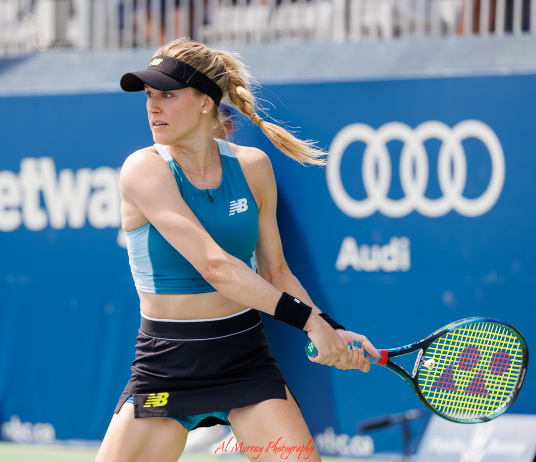 Female tennis player prepares for a backhand shot during a professional match with a blue stadium backdrop and blurred sponsor signage behind her. The crisp action and focused expression reflect the dynamic style of tennis photography around the world.