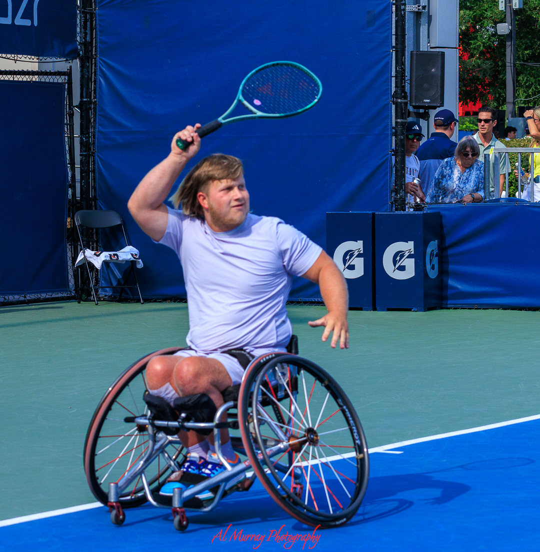 Wheelchair tennis player raises a racket to return a shot during a match on an outdoor blue court while spectators watch from the sidelines. The action focused composition highlights the intensity and athleticism often captured in sport photography in Ontario.