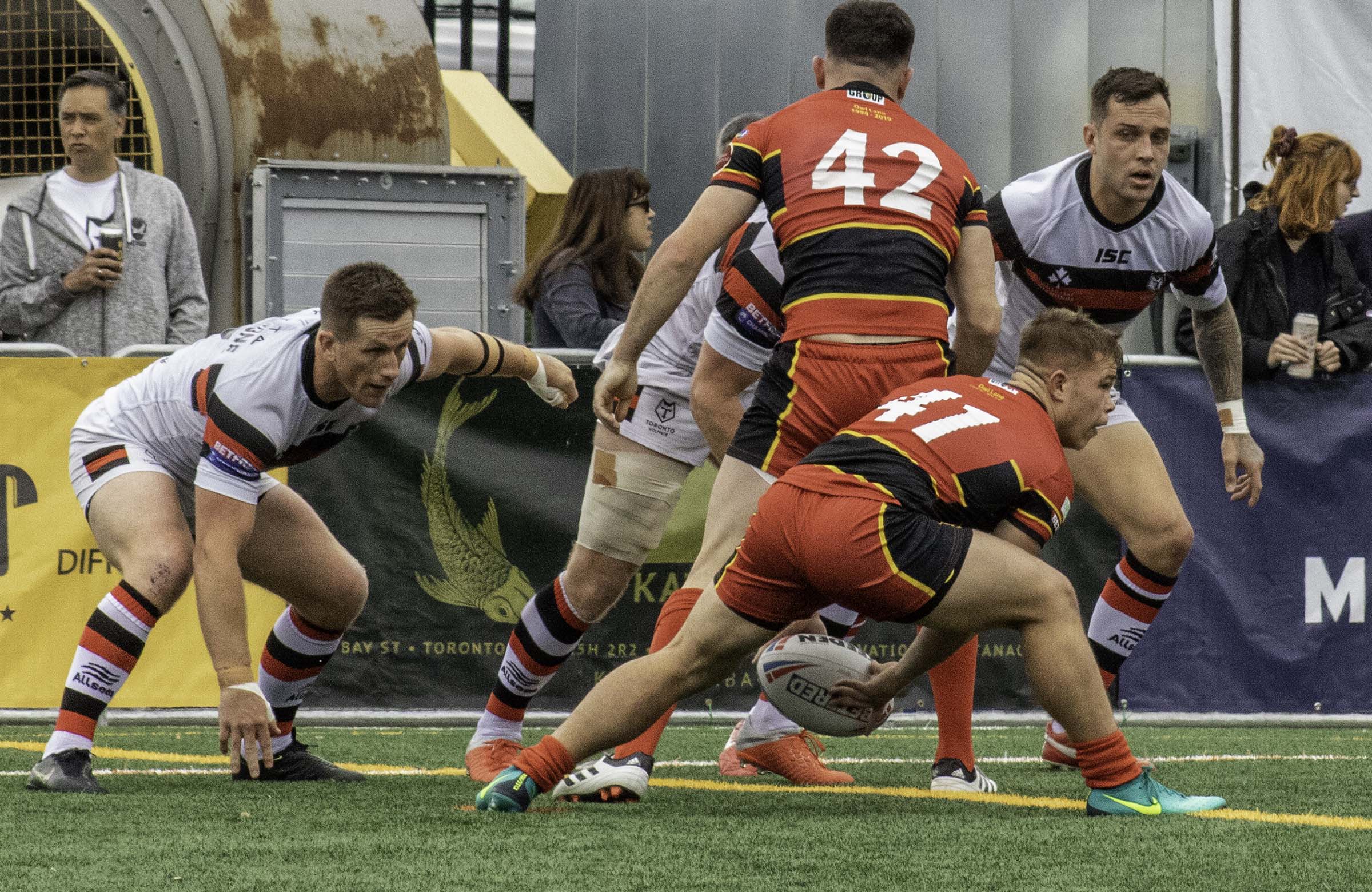 Rugby players in red and black uniforms face off against players in white jerseys during an outdoor match on a turf field. One player crouches with the ball while teammates prepare for the next play, capturing the fast paced action and intensity often documented by a rugby photographer.