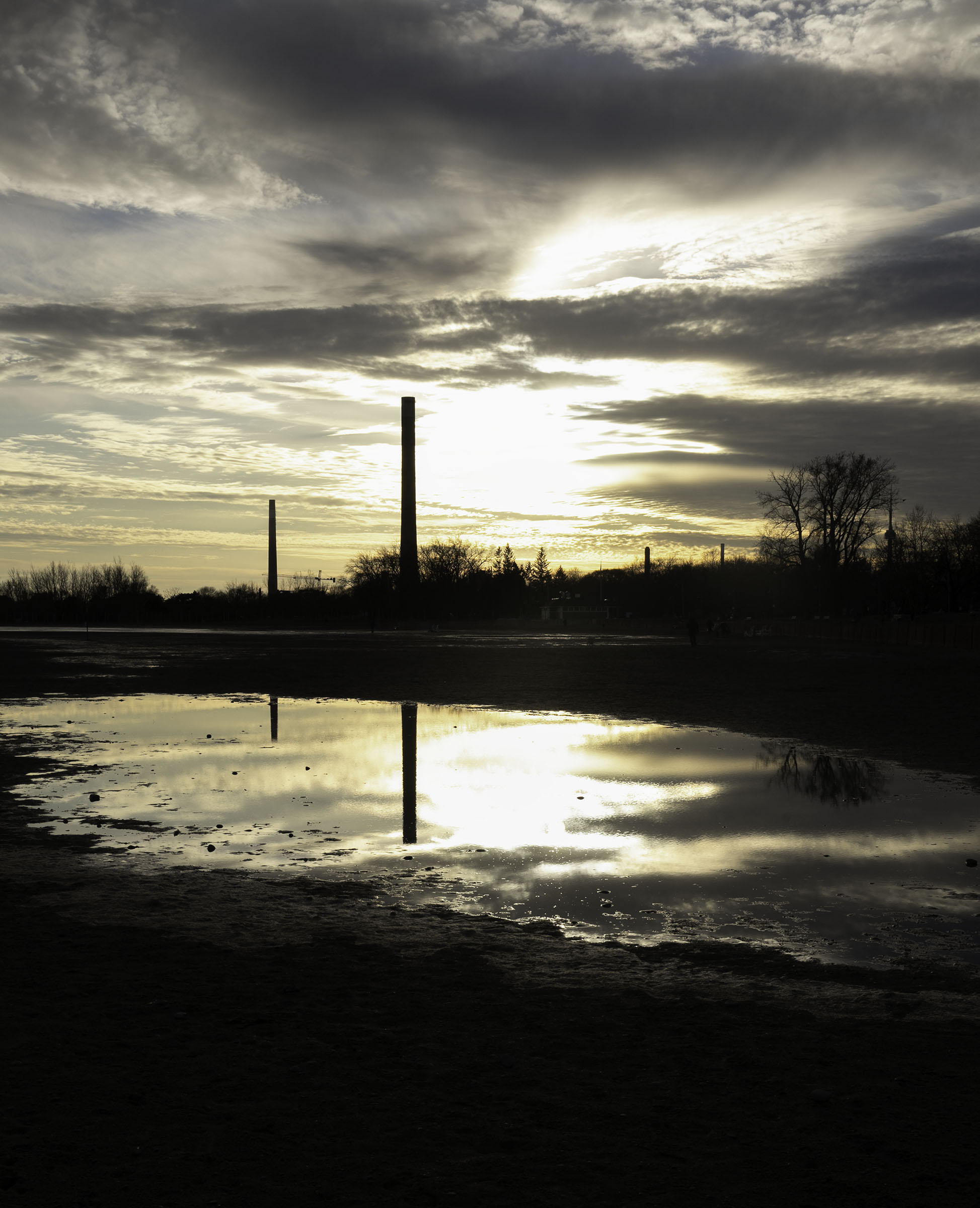 Sunset light breaks through thick clouds above a reflective puddle in an open landscape with silhouetted trees and tall industrial chimneys in the distance. The dramatic sky and mirrored reflections create a moody scene captured through the perspective of a travel photographer.