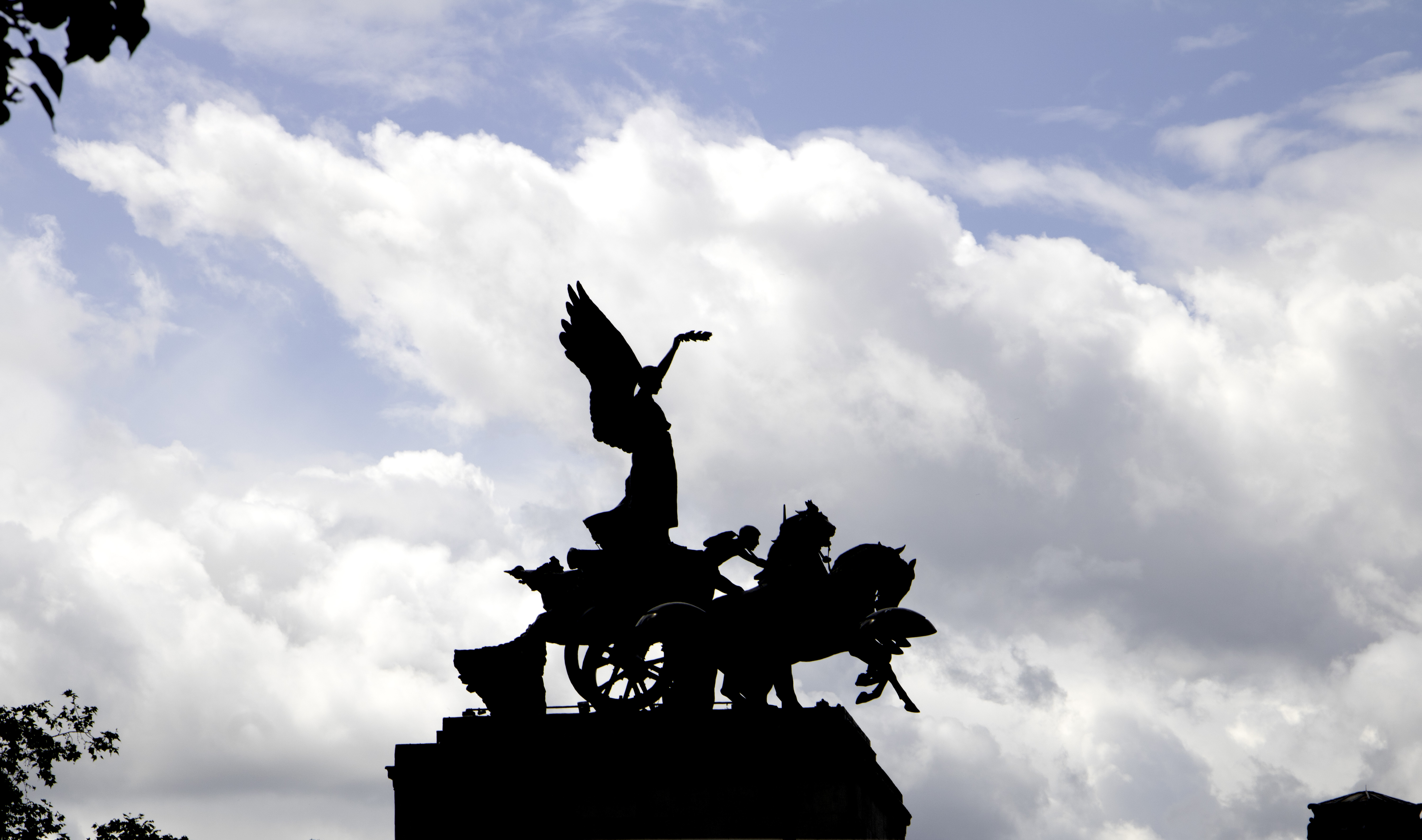 Silhouette of a winged statue standing on a horse drawn chariot against a bright blue sky filled with large white clouds. The dramatic contrast between the dark monument and soft clouds creates a striking scene often associated with travel and landscape photography.