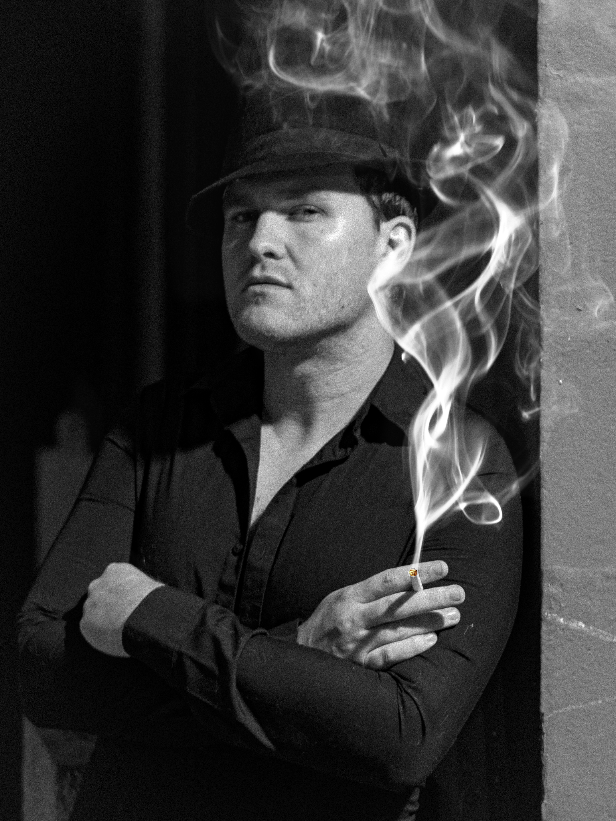 Black and white portrait of a man in a dark shirt and fedora leaning against a wall while holding a cigarette with smoke curling dramatically around his face. The moody lighting and confident pose create a cinematic style often featured in branding and portrait photography in Woodstock.